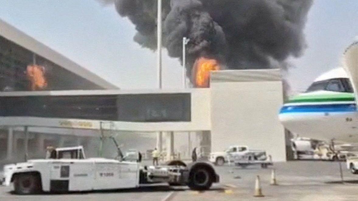 Una columna de humo negro se eleva sobre un edificio de un aeropuerto, con llamas anaranjadas visibles en la fachada. En primer plano hay un vehículo de remolque de aeronaves y, a la derecha, parte de un avión con franjas verdes y azules.
